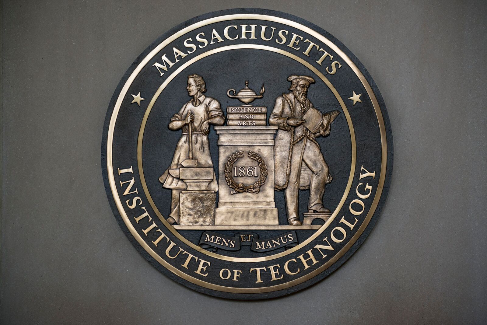 A large, metal rendering of the MIT seal. The background is dark gray, and the type and illustration are in a bronze tone.