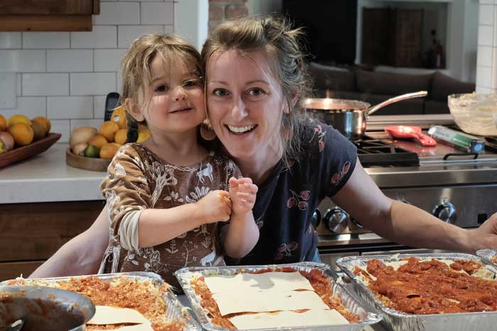 Rhiannon Menn smiles with her toddler in the kitchen, with big trays of lasagna in the foreground